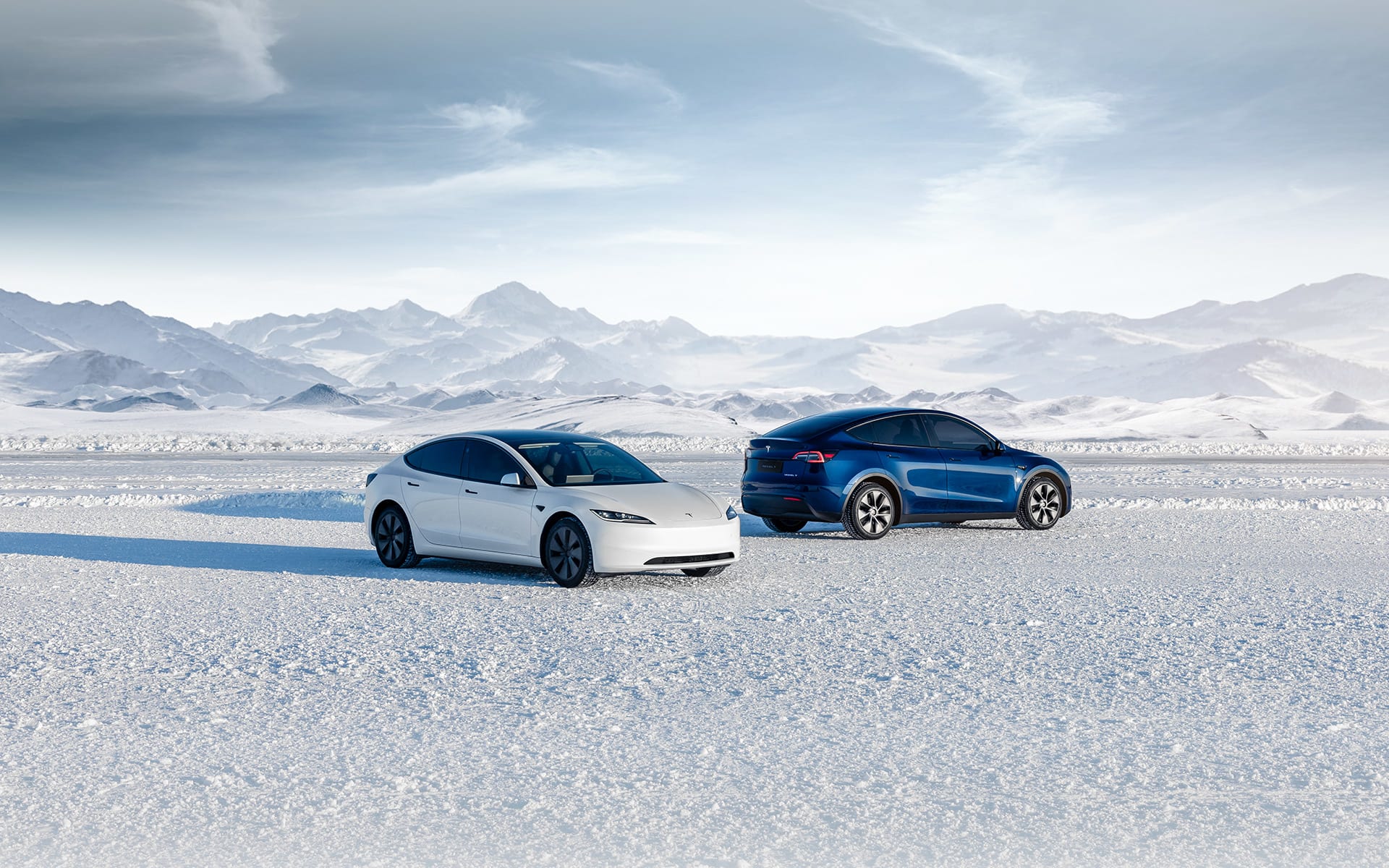 A white Model 3 and a blue Model Y parked on a snowy plain in front of snow-covered mountains
