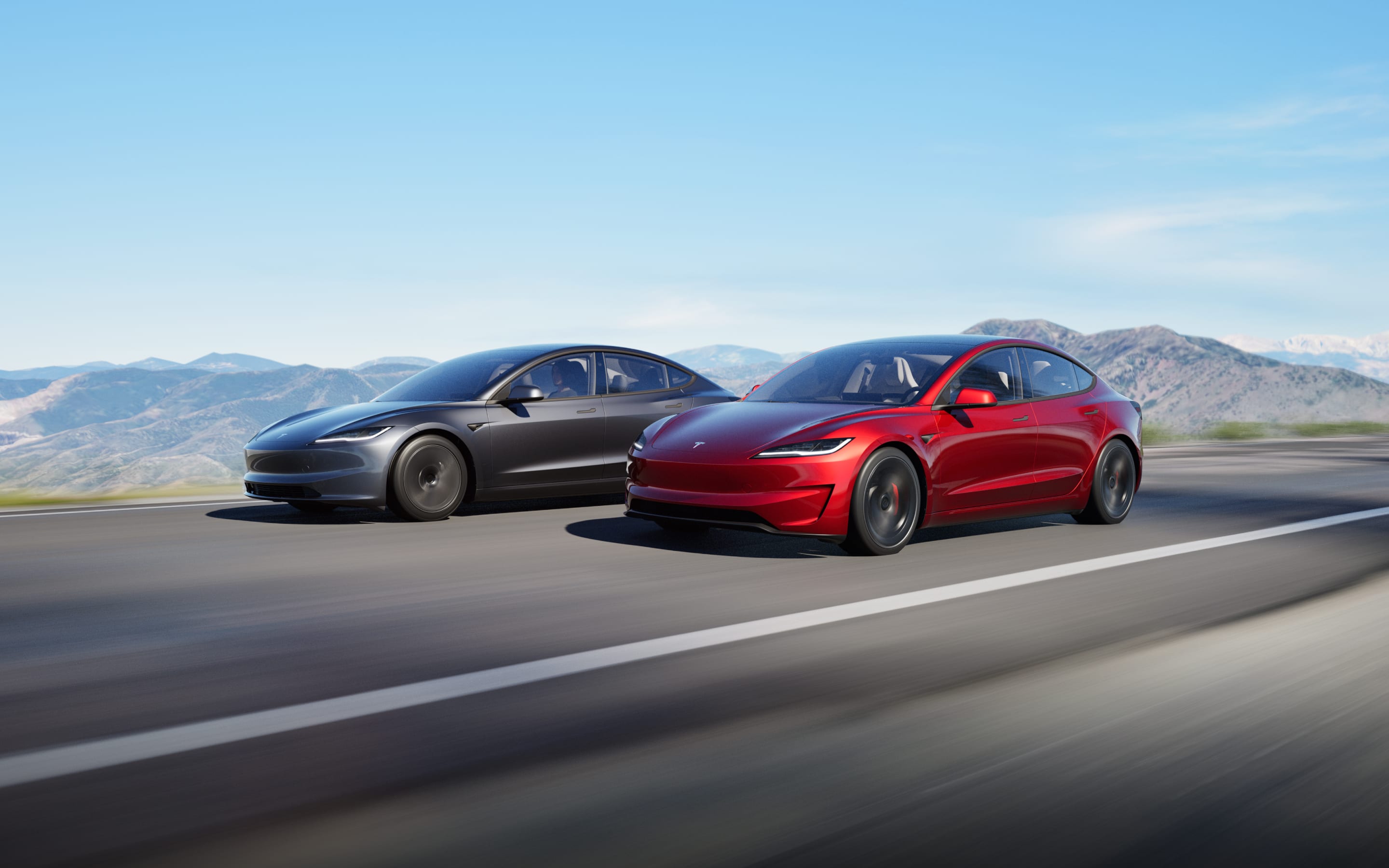 A red Model 3 driving alongside a grey Model 3 on the road with mountains in the background