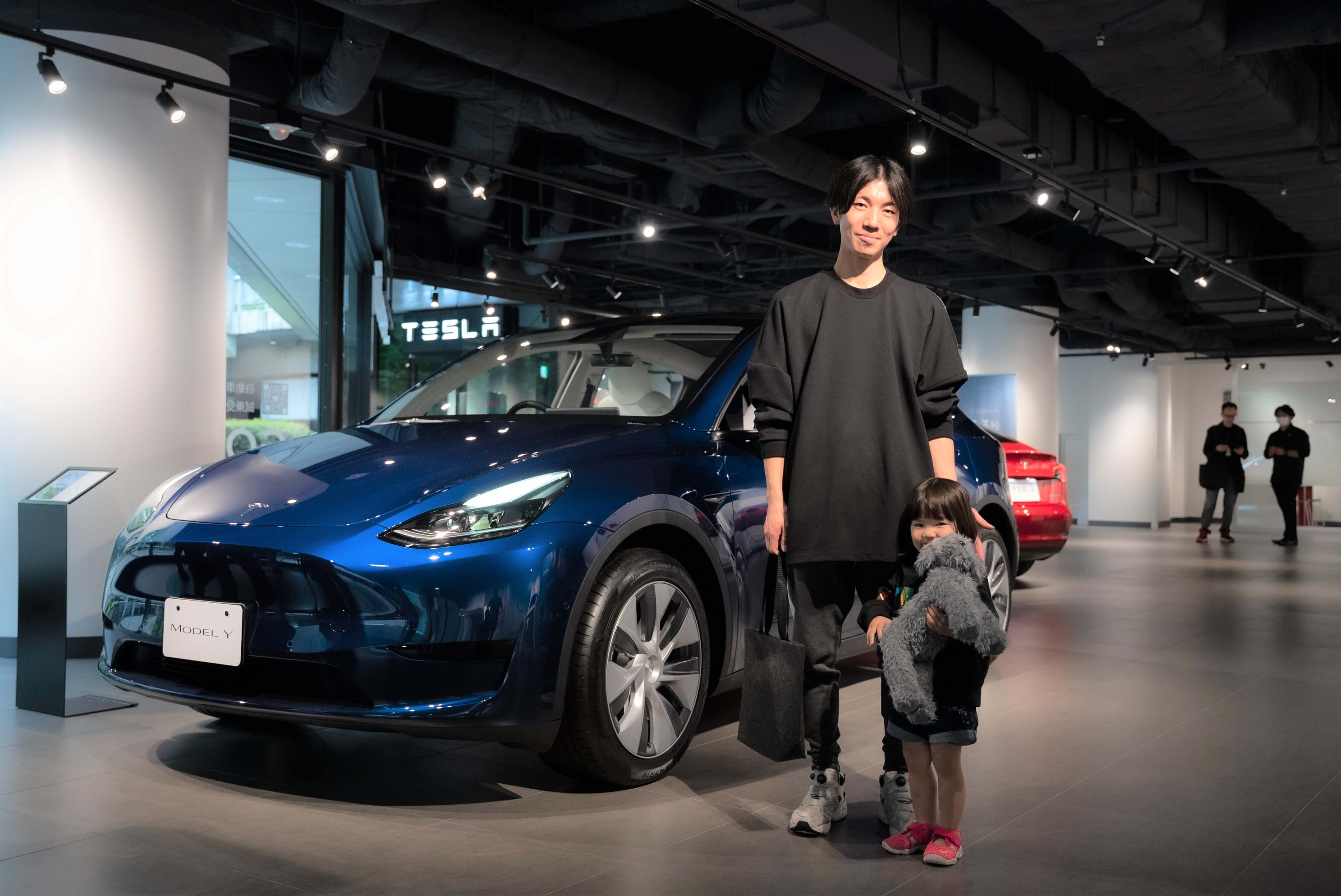 A Japanese Tesla owner with his daughter at the front of a blue Model Y at Tesla Shinjuku store