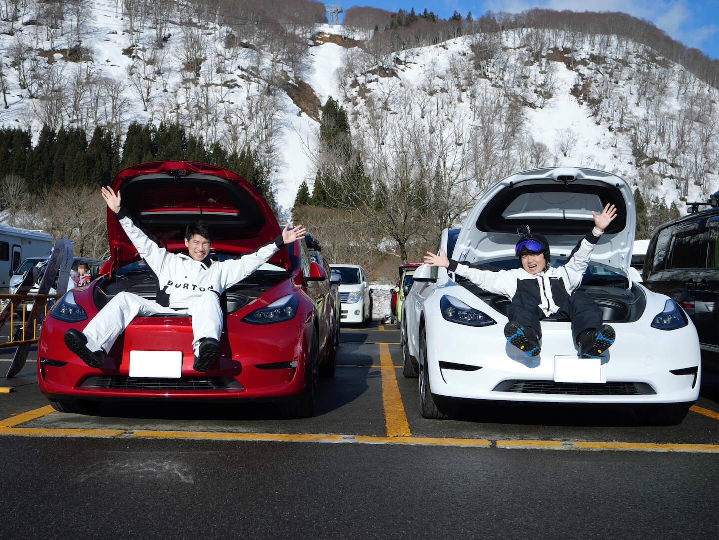 Tesla owners, Daiki and Ray, sitting on the front trunk of a red Model Y and white Model Y at a ski parking lot