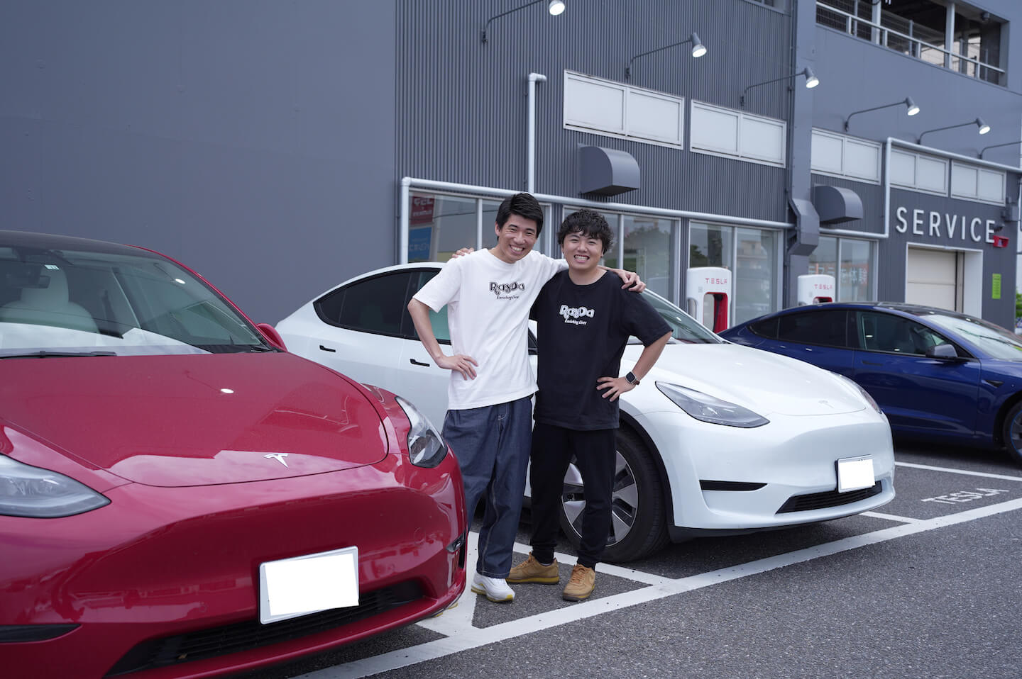 Tesla ownners, Ray and Daiki, standing next to their red Model Y and white Model Y at the parking lot of Tesla Service Cente
