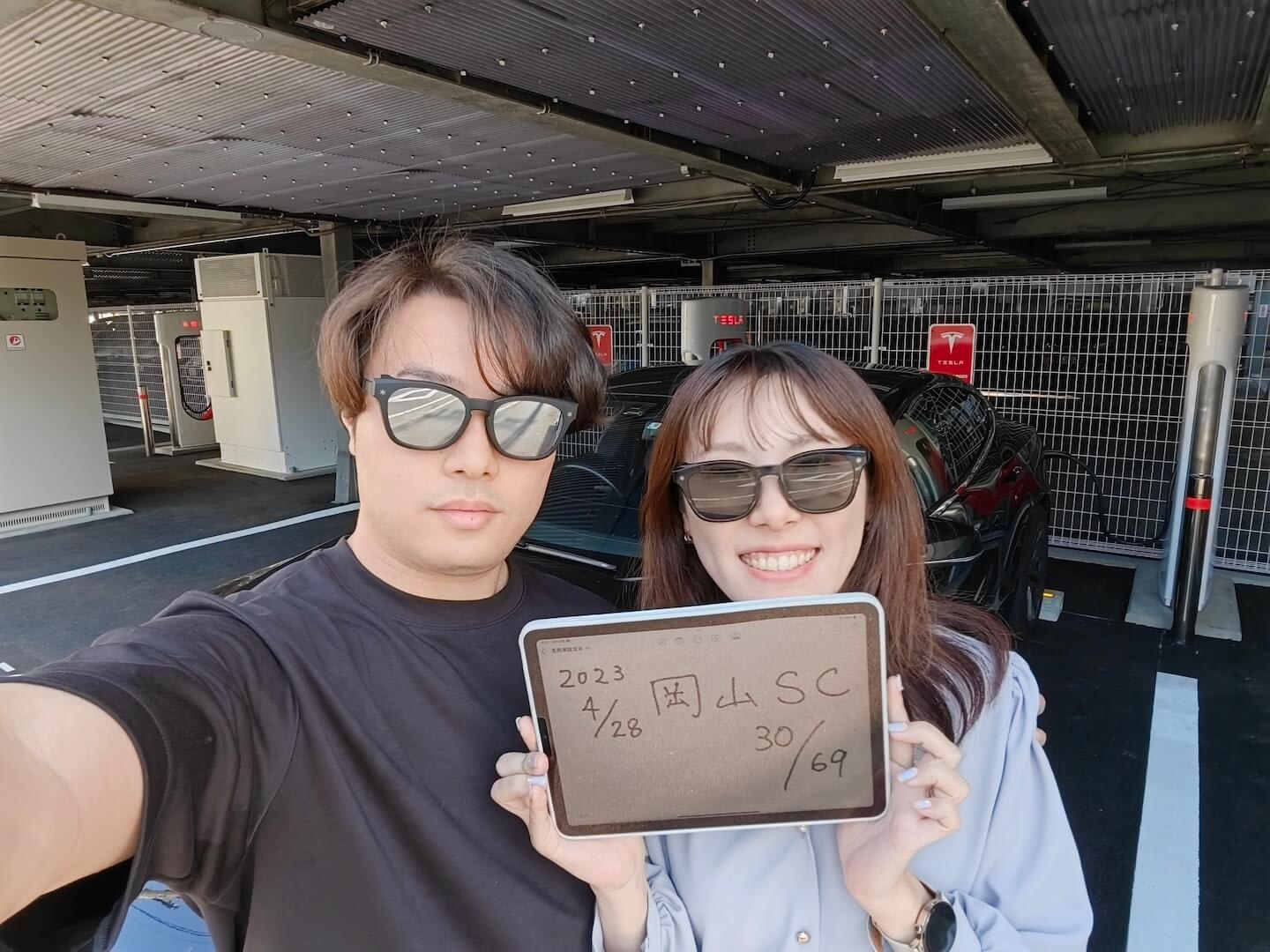 Tesla owner couple with a tablet displaying "2023/4/28 Okayama Supercharger 30/69" while charging their black Model S