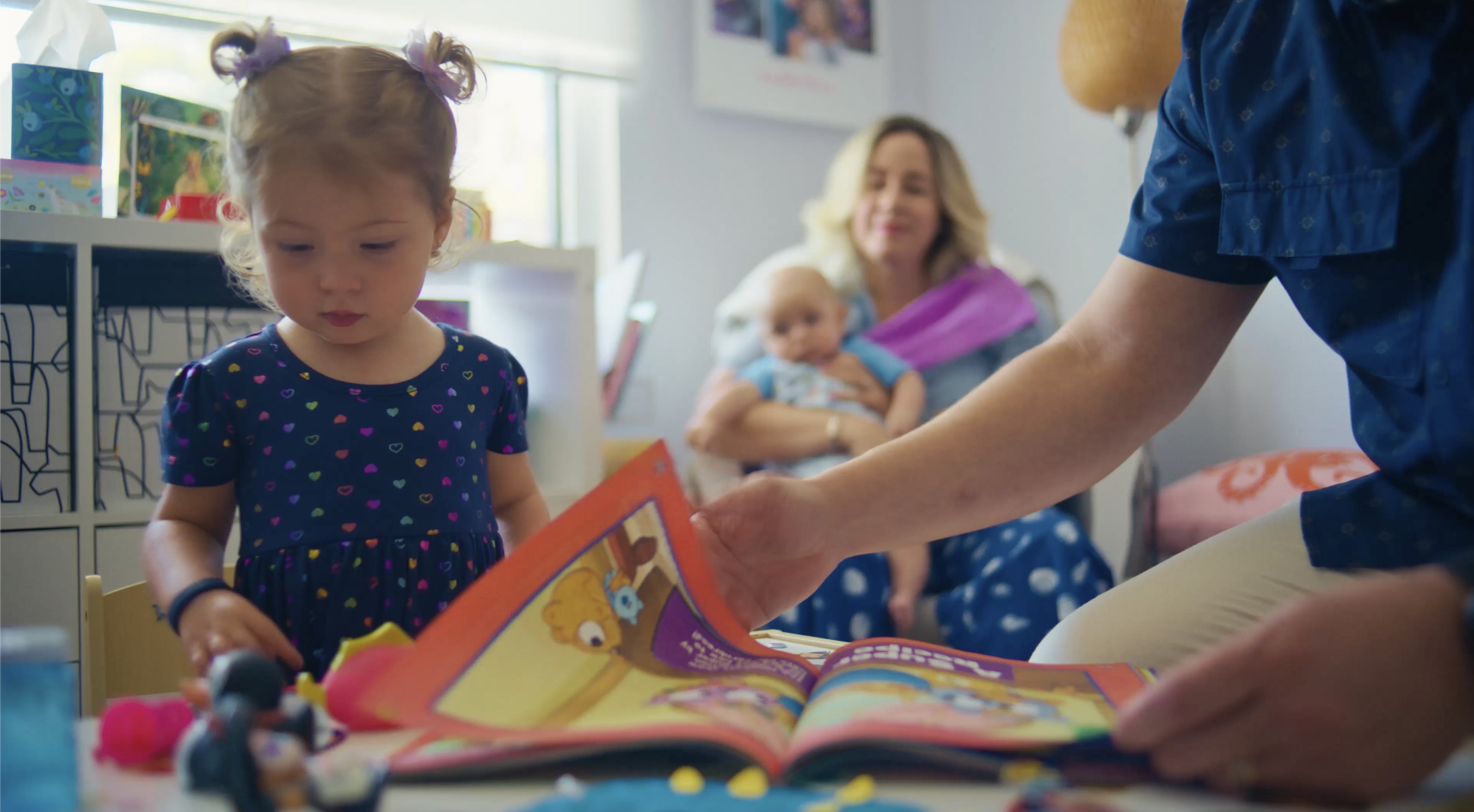Father reading a book with a young child while mother holds a baby in the background