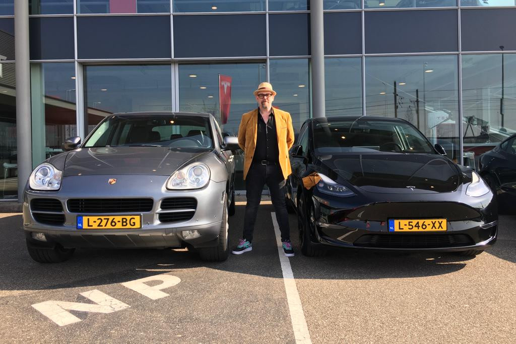 Man staat tussen een Solid Black Model Y Long Range en een Porsche op een parkeerplaats.