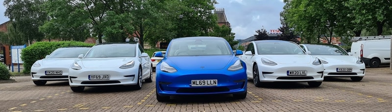 Four white Model 3's and one blue Model 3 in a parking lot
