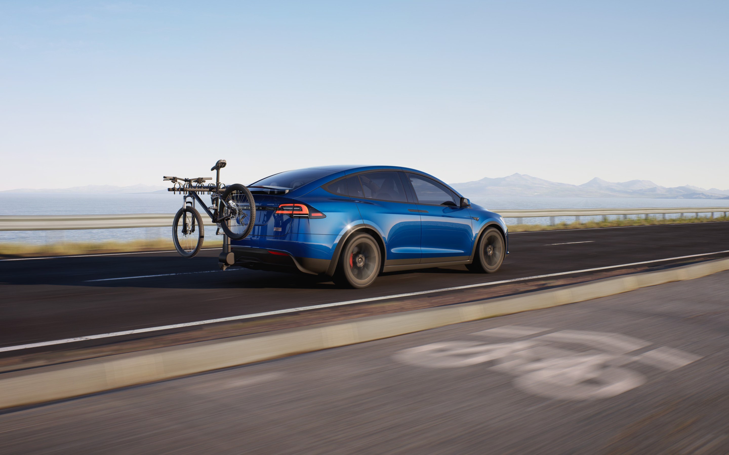 Blå Model X Plaid set bagfra, der kommer kørende på en landevej med en cykel monteret på et cykelstativ